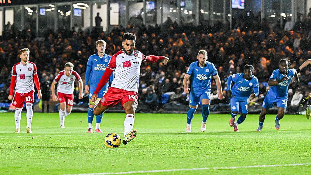 Jamie Reid wearing a red and white Stevenage kit kicks the yellow and black football. In the background there are two more Stevenage players and four Peterborough United players visible on the pitch in front of a stand of fans. 
