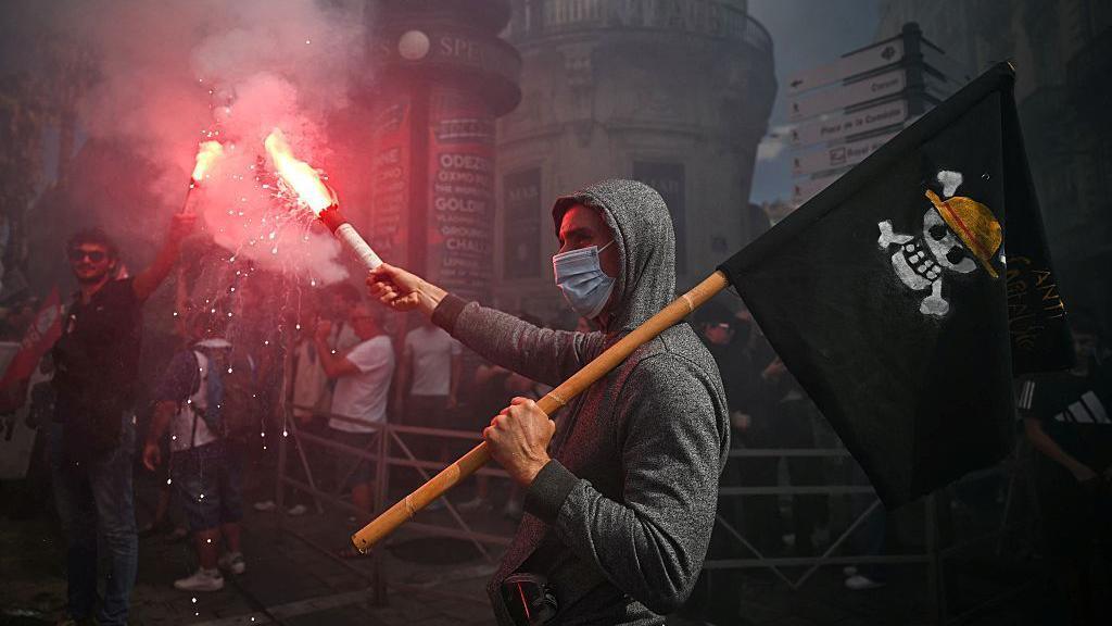 A man holds a flare at a protest in Paris and brandishes a black flag with a skull