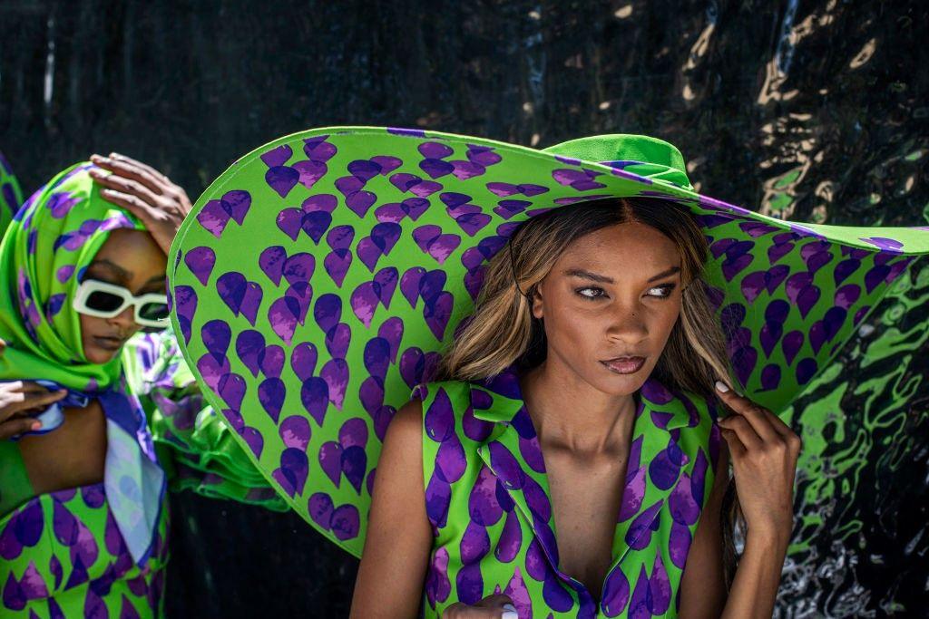 Models wait backstage wearing huge hats and clothing featuring the same green-and-purple print.