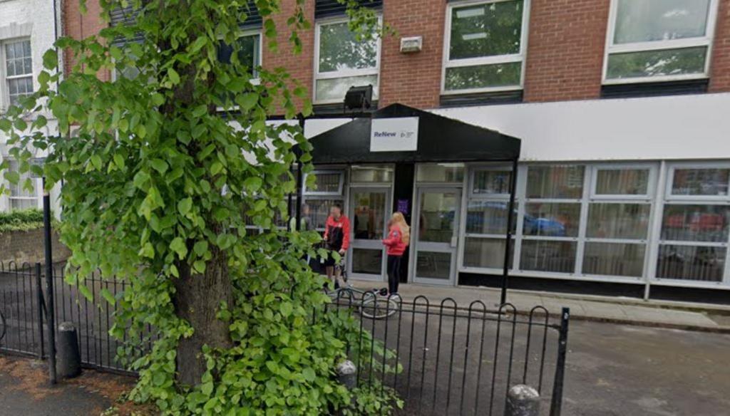 View of the building with people standing outside the entrance which sits behind a black metal railing 