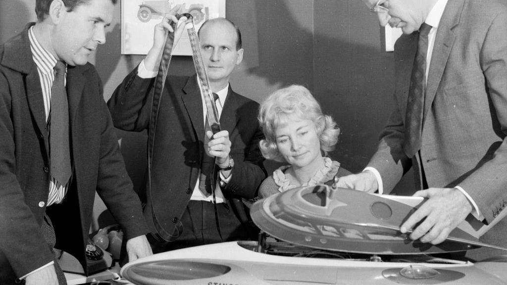 A black and white photo of three men in suits and one woman examining a plastic model of a 'Stingray' submarine.