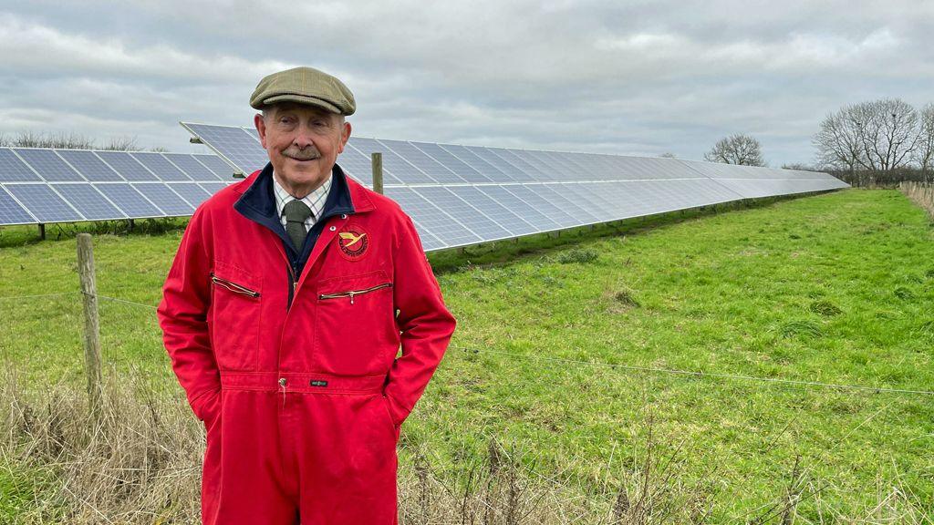 William Rose pictured in front of one of his fields, which hold a small number of solar panels. He is wearing a shirt and green tie, with a red boiler suit and a flat tweed cap. His hands are in his pockets. Behind him is a field, with grass, and two lines of metal solar panels. It is a cloudy day.