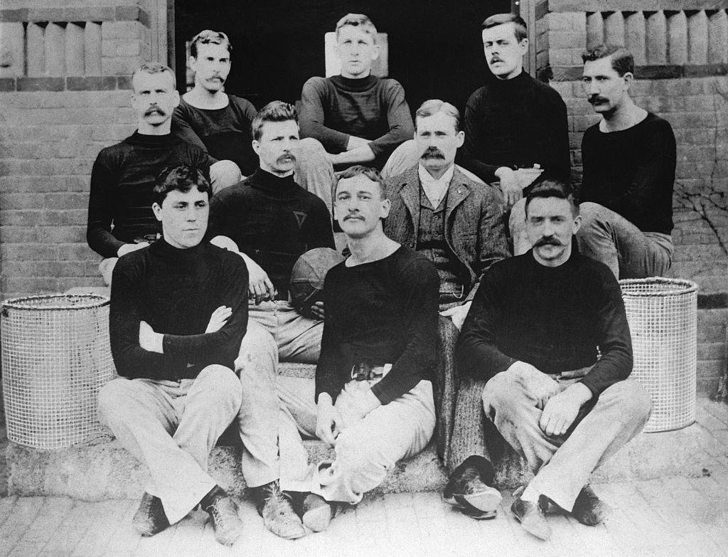 The world's first basketball team - inventor and coach James Naismith with nine players at Springfield College Gymnasium in 1891. Naismith is in a suit, while the players each wear dark, long-sleeved tops with round necks and light trousers. Alongside the players, who are posing in a group shot, are two baskets.