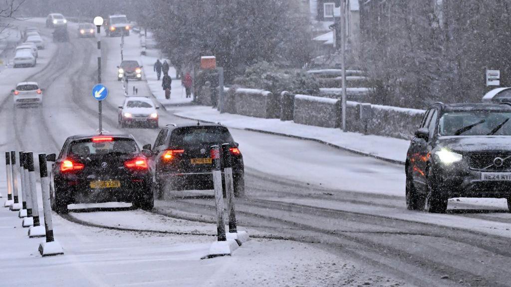 Cars driving on a road with snow falling and starting to settle on the pavement and partially the road itself