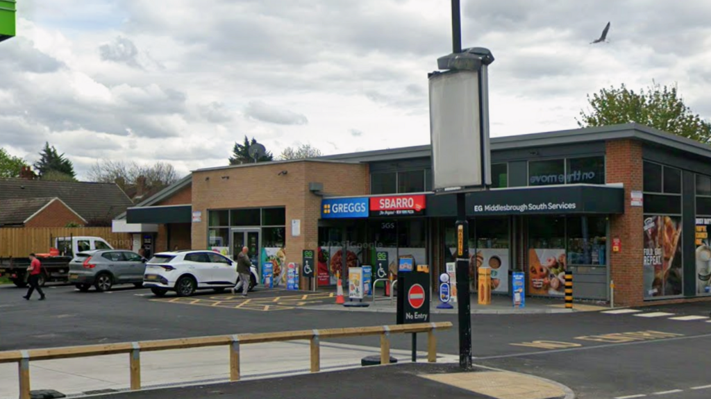 A Google Street View screenshot of the shops at Roseberry Service Station in Middlesbrough. The low, flat-roofed building has signs for Greggs and Sbarro. Two cars and a small flat-bed lorry are parked in front, facing the building and a few people are walking to or from it.