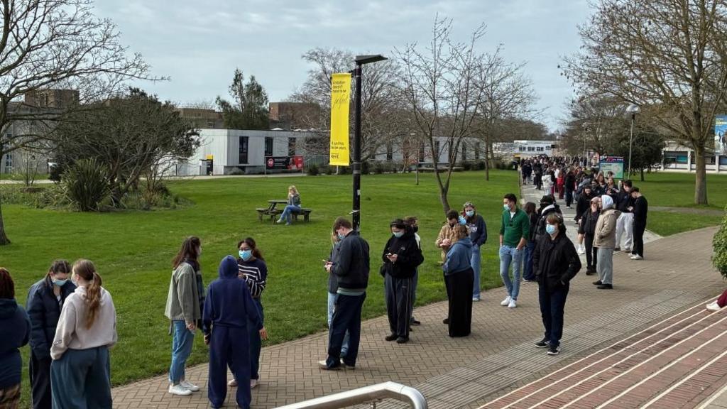 Students (wearing face masks) form a queue on campus as they wait in line to receive antibiotics