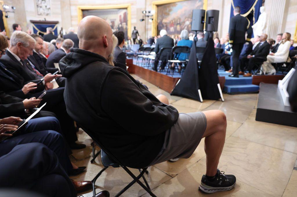 A man in a hoodie, basketball shorts and sneakers sits in the Capitol Rotunda surrounded by men wearing suits