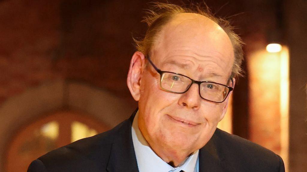 Ken Reid, who is balding and wearing black-rimmed glasses, looks toward the camera wearing a black suit and light blue shirt