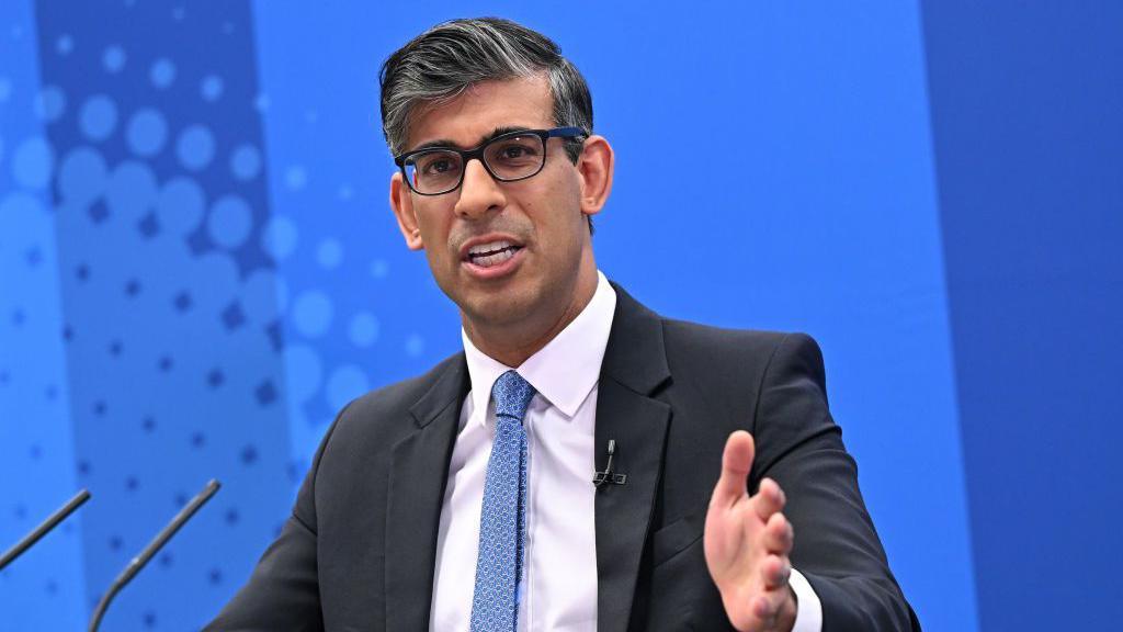 Rishi Sunak speaking in front of a microphone in front of a blue background