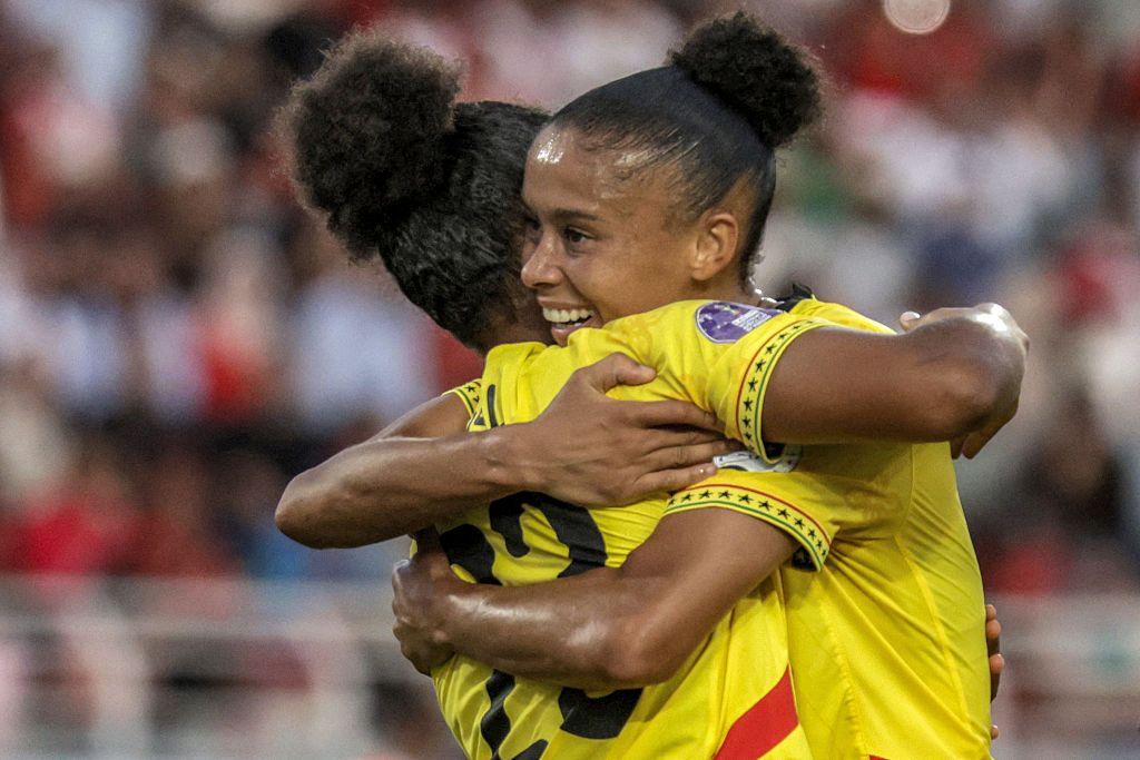 Two women wearing matching yellow football jersey's hug on the pitch.