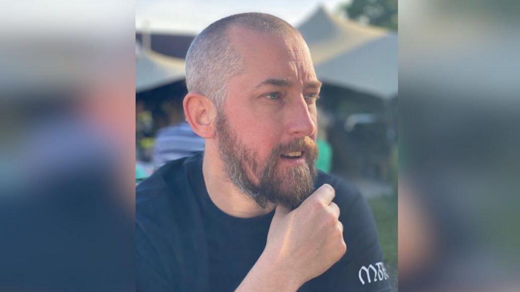 PC Mathew Parker is sitting with his hand on his chin. He is wearing a black T-shirt with white writing on the sleeve. He has a beard and cropped hair. 