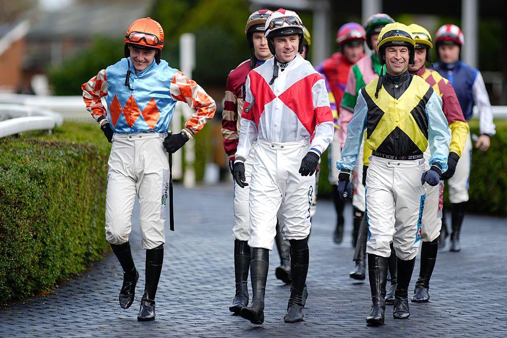A group of jockeys walks along a paved path toward the parade ring, wearing colorful racing silks with bold patterns, white riding pants, tall black boots, and helmets. Green hedges line the walkway. 