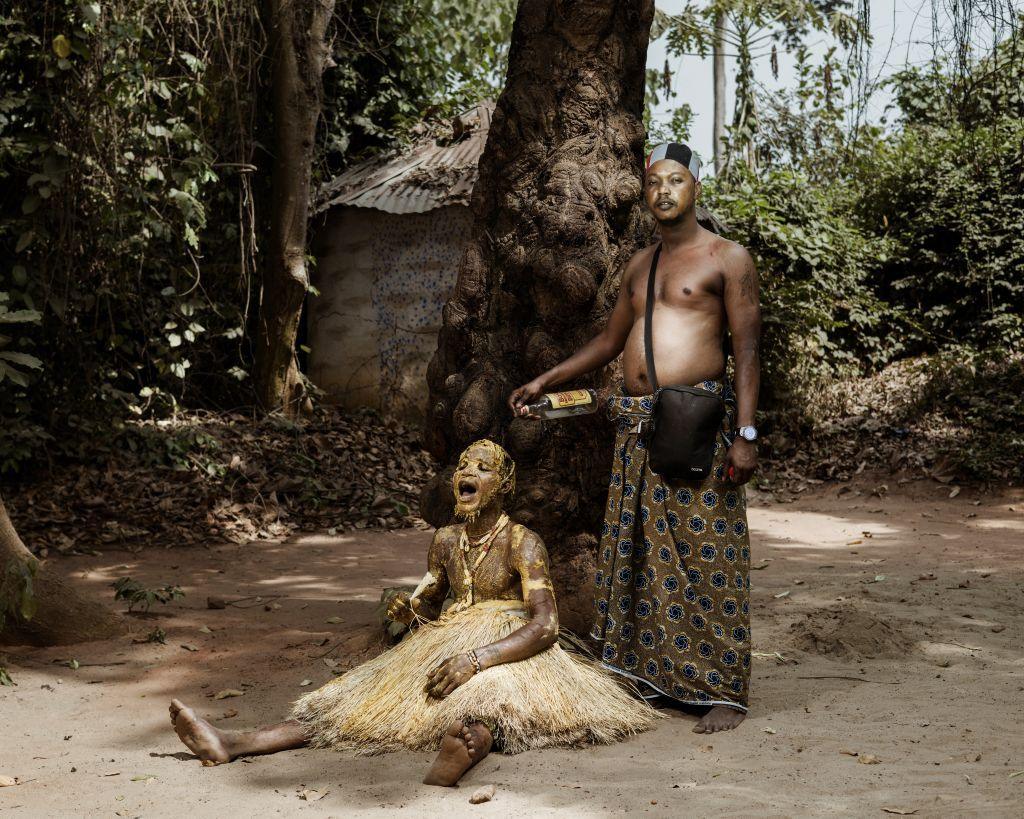 A man, standing, drizzles gin over a young person sitting on the floor, who is covered in red palm oil mixed with maize flour, giving it a golden hue at a ceremony at the at the annual Voodoo festival in Benin, Ouidah - Thursday 9 January 2025.