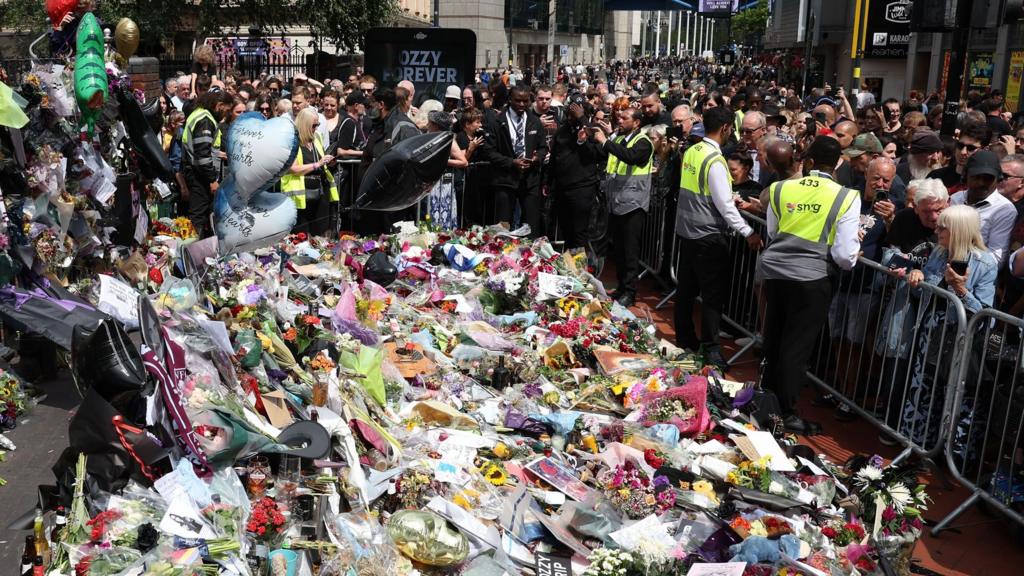 An area covered deeply in tribute ballooms and flowers, surrounded by crowd barries and hi-viz-wearing security guards. Behind, a crowd stretches as far as the eye can see along the road behind.