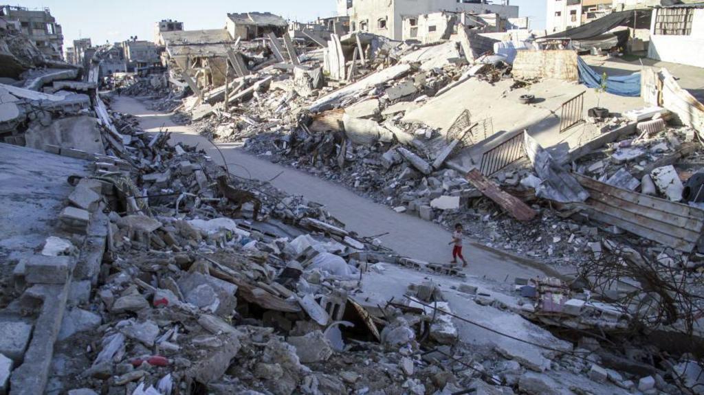 A wide shot of Jabalia, where a child stands on a road where either side of him is grey rubble.