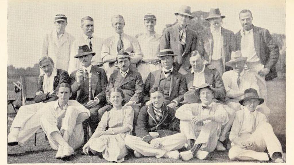 A black and white photo of a group of men and women in a team photo on a cricket pitch.
