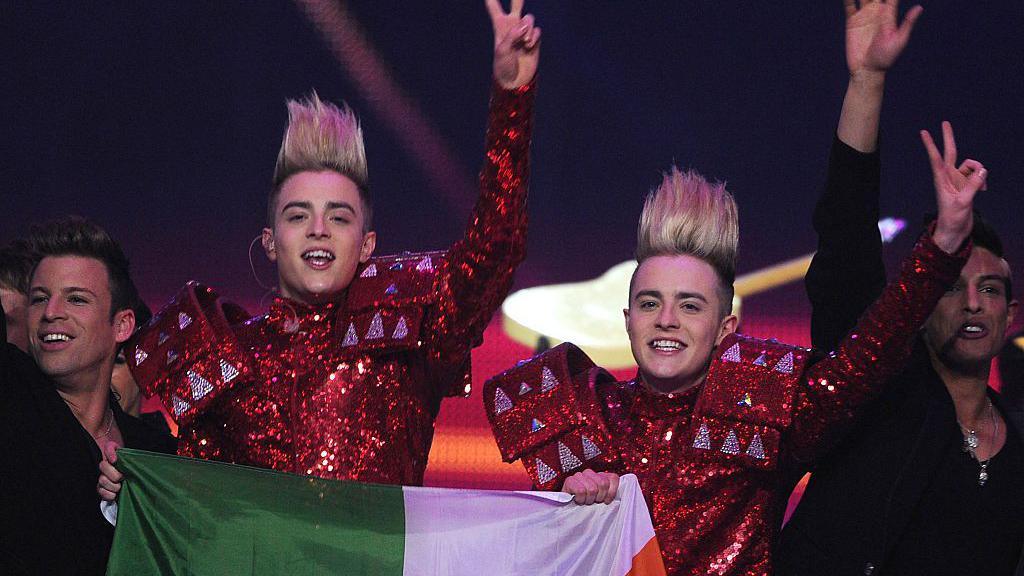 Two men with blonde hair wearing bright red suits holding an Irish flag. 