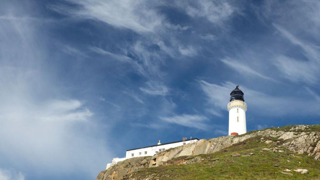 A lighthouse building on a rocky outcrop sticks out against a blue and whispy clouded sky