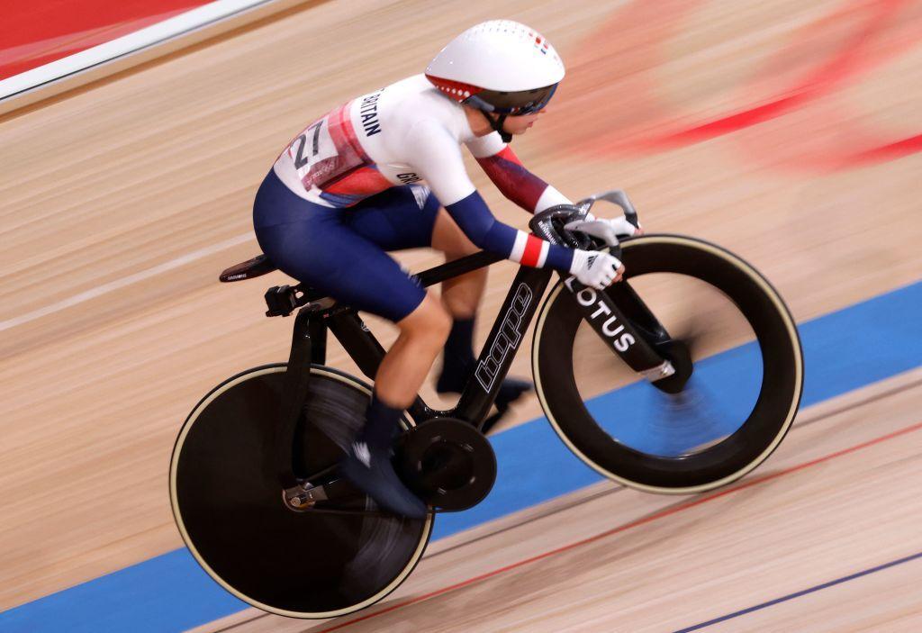 Laura Kenny wears a blue and white team GB lycra bodysuit and white helmet, clipped into a black racing bike as she hurtles round a velodrome track.