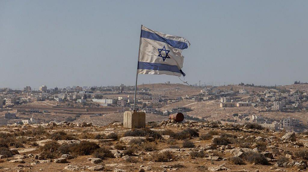 An Israeli flag is planted into the ground, in a barren, hilly landscape in the occupied West Bank