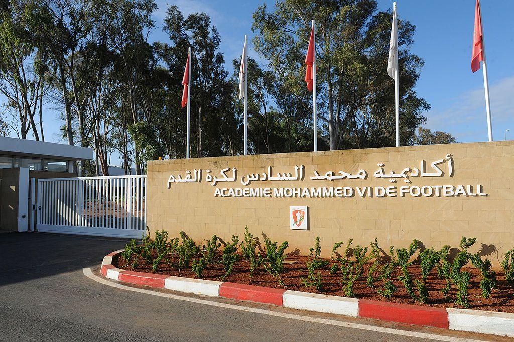 The outer wall of the Mohammed VI Football Academy in Morocco is shown with a white coloured gate and sand-coloured wall. There is a flowerbed close to the wall, white lettering in English and Arabic on the wall, and red and white flags hanging limp on white flagpoles. There are tall green trees behind the wall against the backdrop of a blue sky