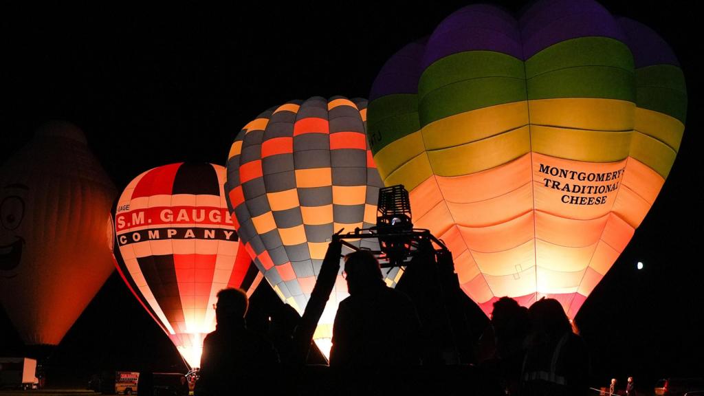Several balloons lined up in the dark with burners on, lighting the balloons up