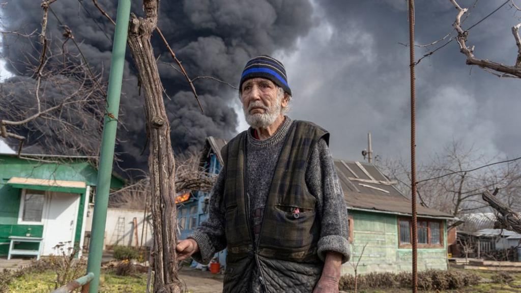 A local resident stands in the courtyard of his house while smoke rises from a fire following a strike on the outskirts of Odessa on March 11, 2025, amid the Russian invasion of Ukraine