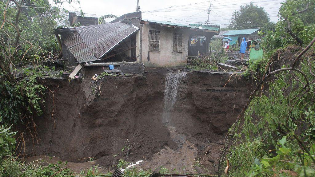 A man in a blue rain poncho looks at a landslide-hit residential area surrounded by greenery and trees in Guinobatan town, Albay province, south of Manila. A small concrete house, with a tin roof collapsed beside it, sits on the edge of a precipice that has been stripped to dirt by a landslide, with water running off it.