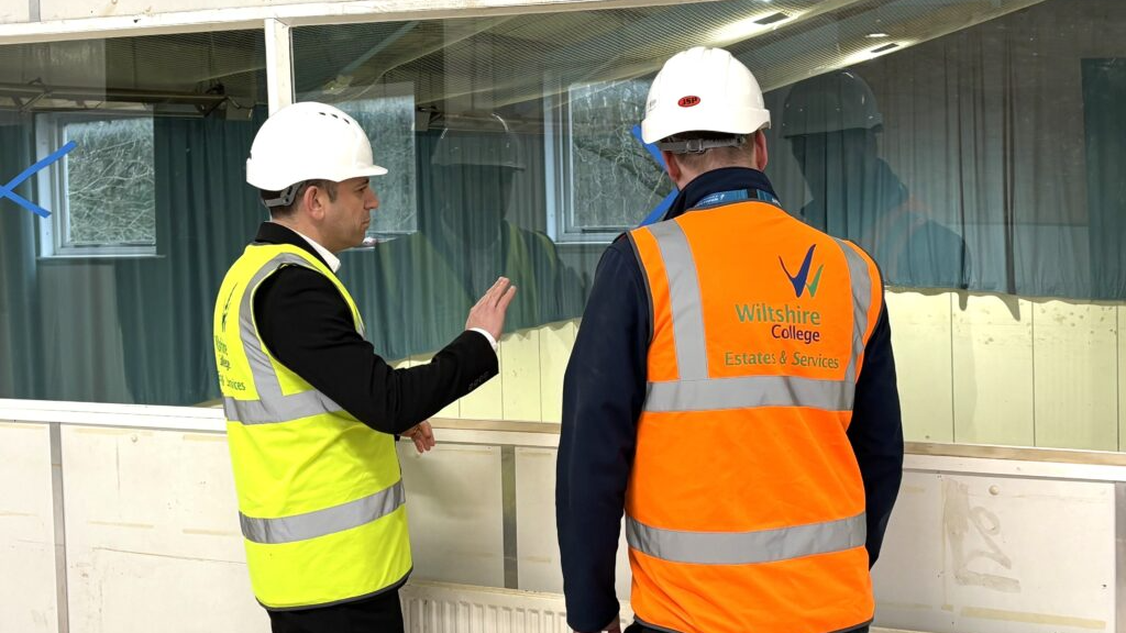 Two men in high-vis vests are standing with their backs to the photographer and looking through a window. They are both wearing white hard hats.