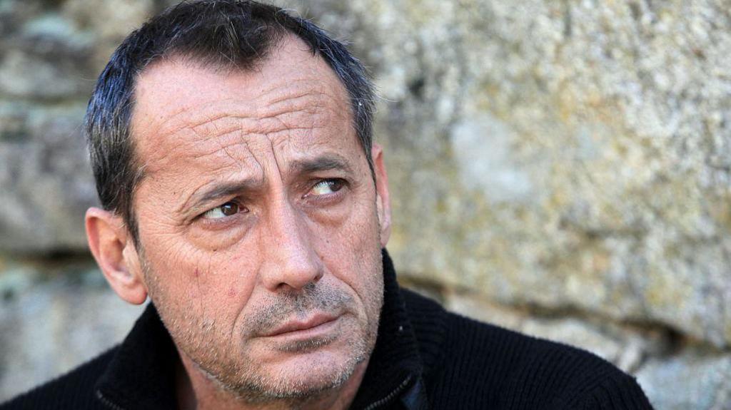 A man in a black sweater looks askance with a short beard and greying hair