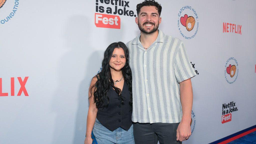 Kayla Vesia, wearing a black blazer and jeans, stands smiling next to husband Alex Vesia, wearing a button-down and jeans, in front of a Netflix step-and-repeat