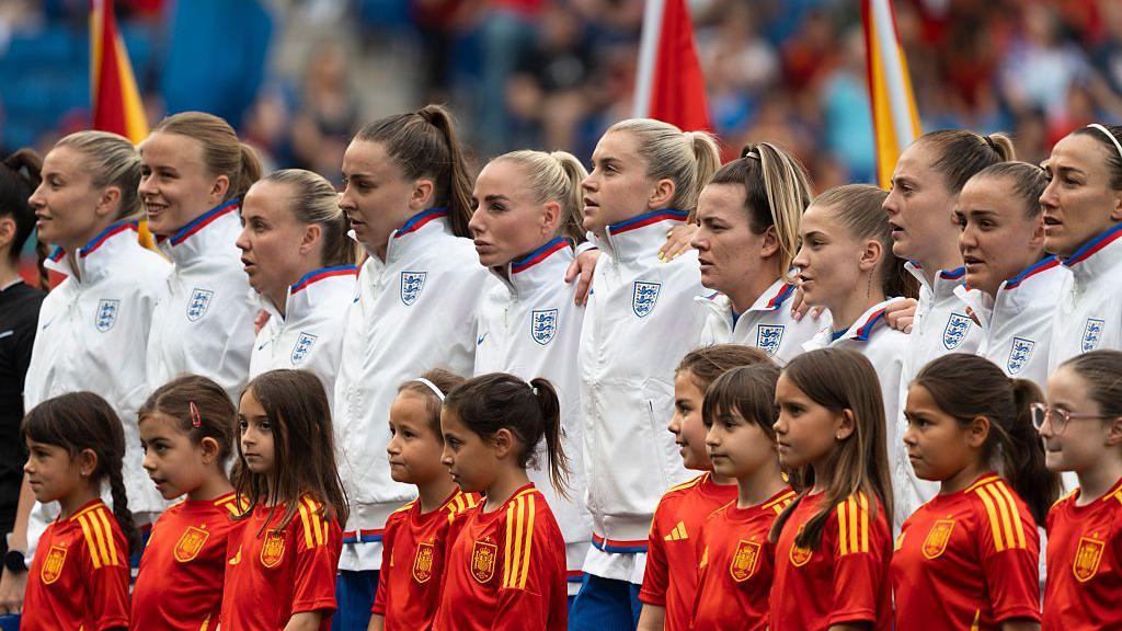 The England national team listens to the national anthem before a football match, with young mascots in front of them