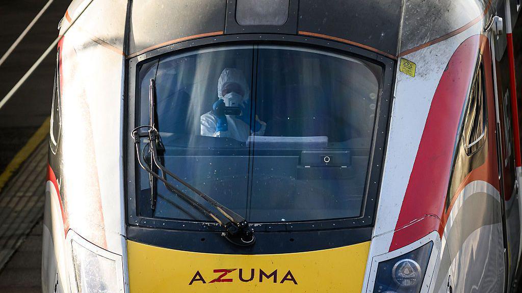 A forensics officer sits in the train driver's seat. They are wearing a protective white cover-all and a white face mask holds a camera up.