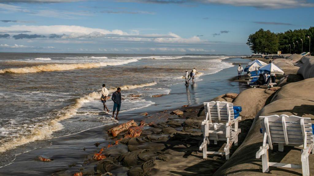 A beach in Bangladesh. Waves are gentle but there's sandbags lined up along the sand.
