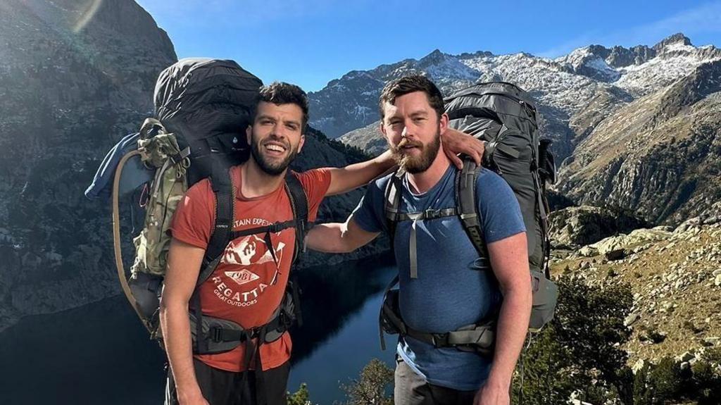 Aziz Ziriat, left, and Samuel Harris in shorts, t-shirt with large backpacks in front of a lake surrounded by mountains