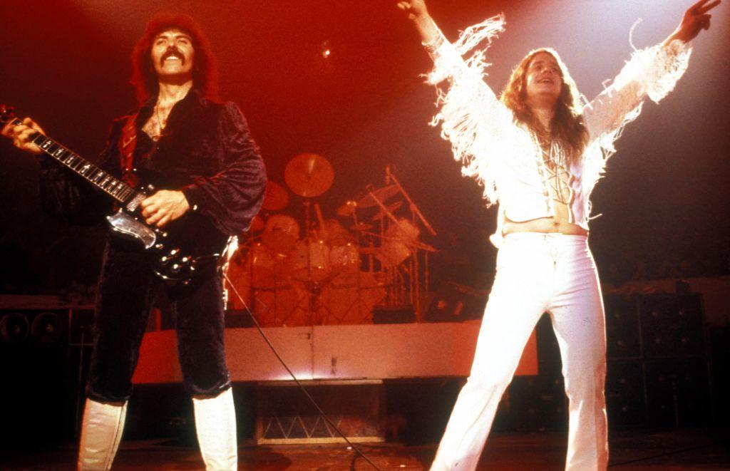 Dark haired guitarist Tony Iommi wearing a black outfit and white boots stands next to lead singer Ozzy Osbourne wearing a white outfit with arms raised