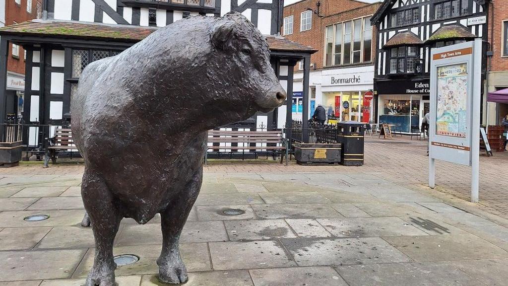 A metal sculpture of a bull, looking to the side, seen on a paved area in a city centre with shops around it, many with Tudor-effect wooden beams separating white panels in the walls.