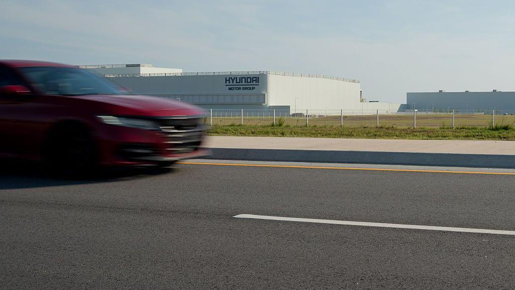 A red car drives by the Hyundai plant in Georgia, a large white building behind a fence in a grassy field