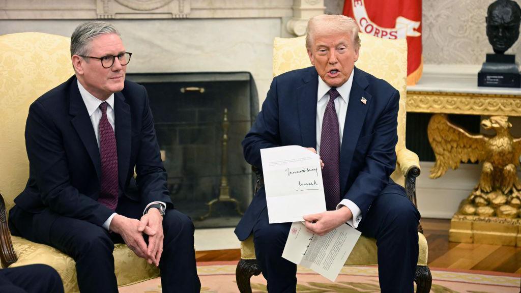 Keir Starmer and Donald Trump sitting together in the Oval Office. President Trump is holding a signed invitation from the King. The two men are dressed almost identically in dark blue suits, white shirts and burgundy ties. 