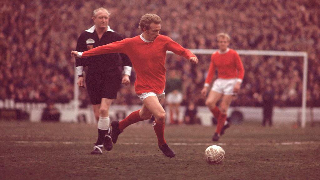 Denis Law aims to kick the ball during a match. He wears a red and white football strip. The referee stands behind him and the stands are full of supporters.