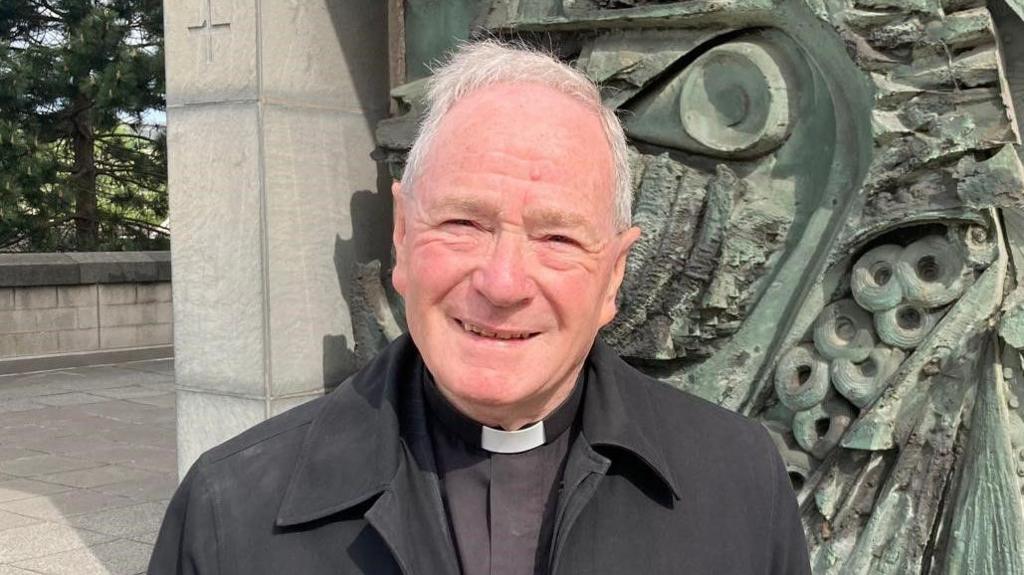 Arthur Siddall, a retired Anglican archdeacon from Garston in Liverpool, has short grey hair. He is photographed wearing a white 'dog collar' and a black shirt under his black coat.