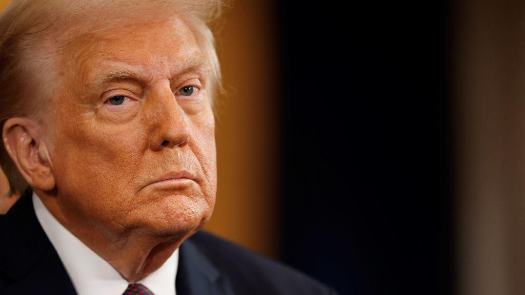 President Trump in close-up. He is on the left of the shot looking just past the camera. He is looking very serious while wearing a white shirt, blue and red dotted tie and dark blue suit.