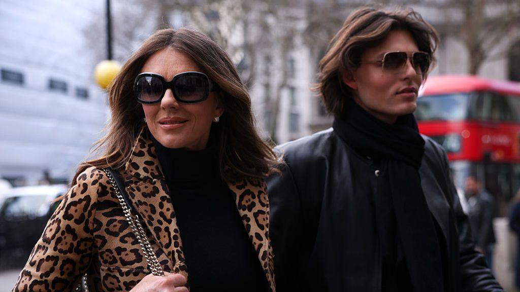 Elizabeth Hurley, me syze dielli dhe një pallto me print leopardi, dhe djali i saj Damian Hurley, gjithashtu me syze dielli dhe një xhaketë lëkure të zezë, mbërrijnë në gjykatë.