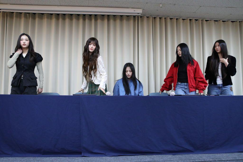 The K-Pop band NewJeans take their seats at a royal blue conference table, ahead of making a statement about the future of their band.