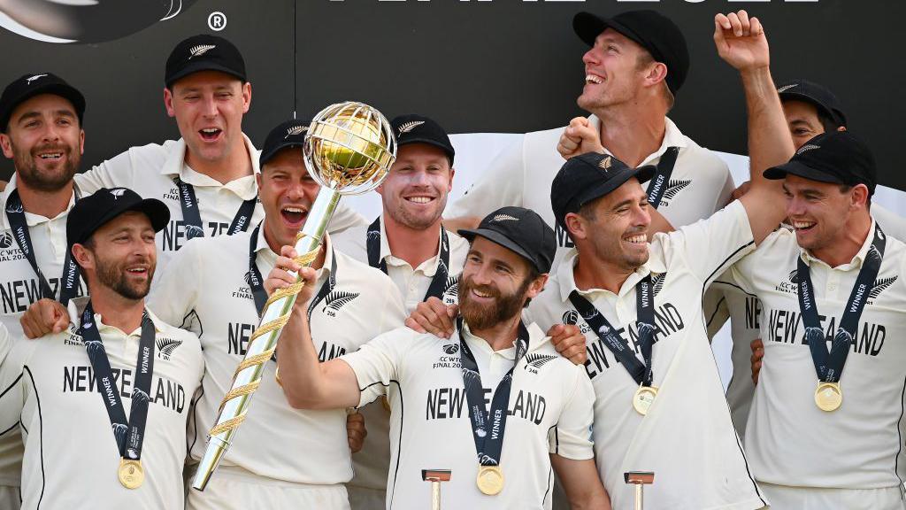 Kane Williamson (centre) holds the silver and gold World Test Championship mace in his right hand and looks at it, smiling, with his teammates around him - all wearing cricket whites with New Zealand in capital letters on the chest, gold medals on black ribbons, and black caps with a white fern on the front