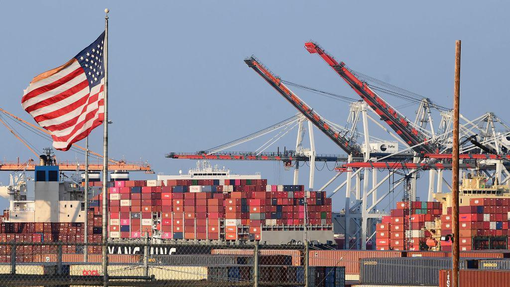 A US flag flies near containers stacked high on a cargo ship at the Port of Los Angeles