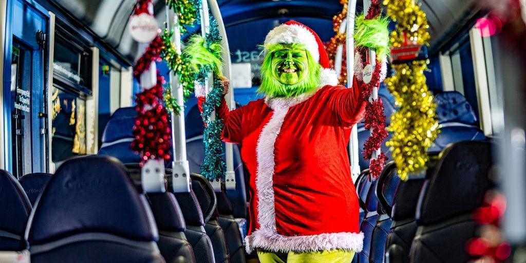 Julie Richardson standing on a bus dressed as the Grinch in a Santa suit. Her face is green and she is smiling.