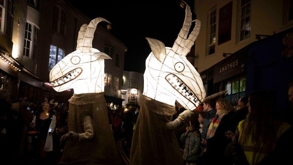 Two lanterns in the shape of creatures with horns and with big grinning mouths showing lots of teeth. There are people lining the streets and buildings are seen either side as well, some lit up and others with lights in upper windows.