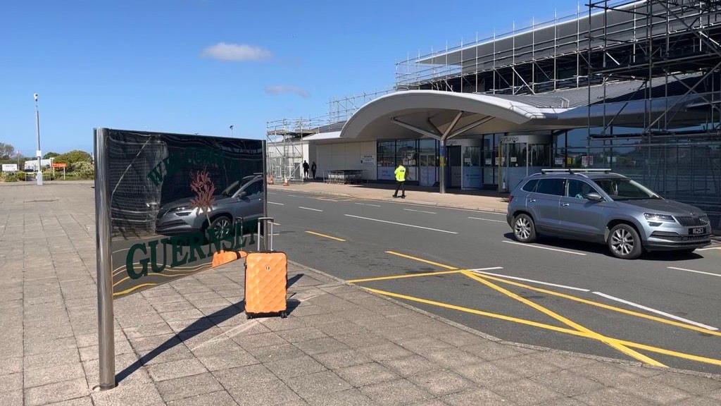 A car drives part a small modern airport. An orange suitcase sits in front of shiny Welcome to Guernsey sign.