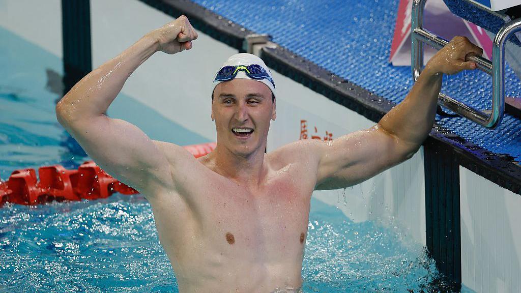 Cameron McEvoy raises his right arm in celebration after setting a new world record. He is standing in the pool and wearing a swimming cap.